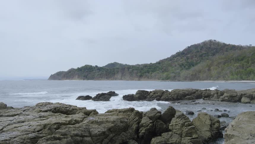 Waves crash onto shore between rocks with forested hills in background
