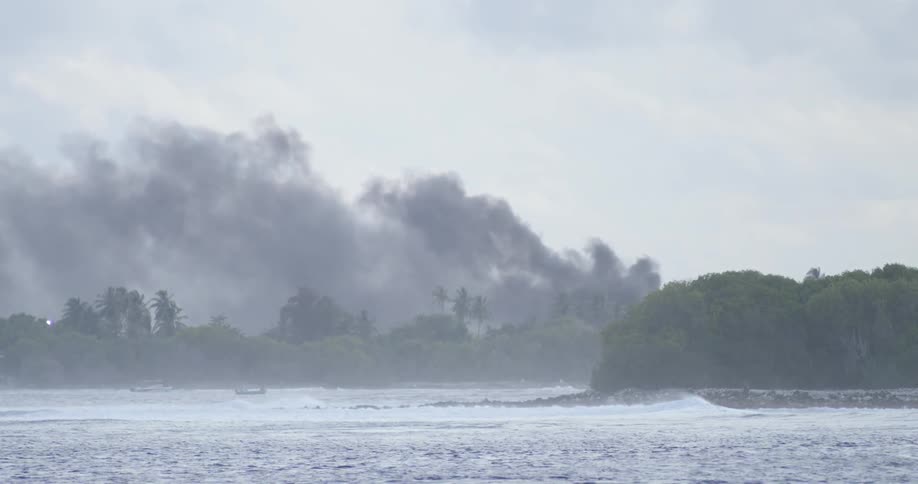 Dark smoke rises into the sky along a coastline