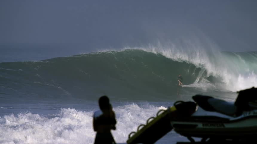 The woman standing on the shore watching a person surf on a wave