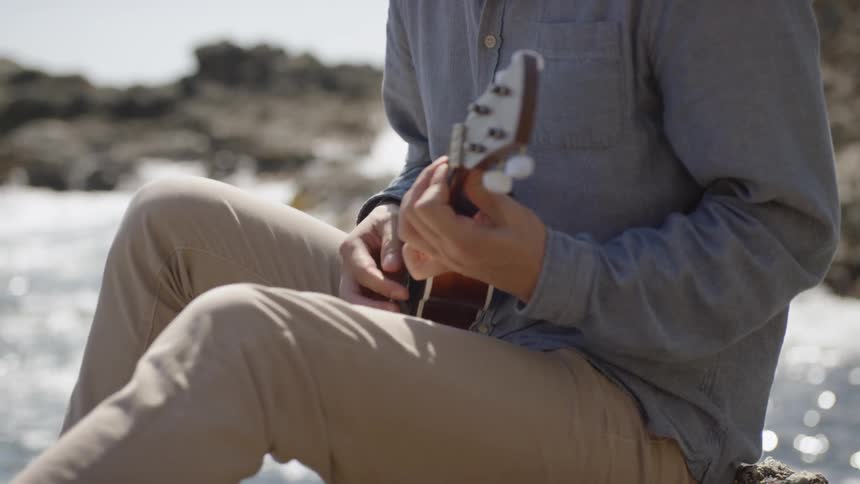 A man sits along a rocky waterway playing a small guitar