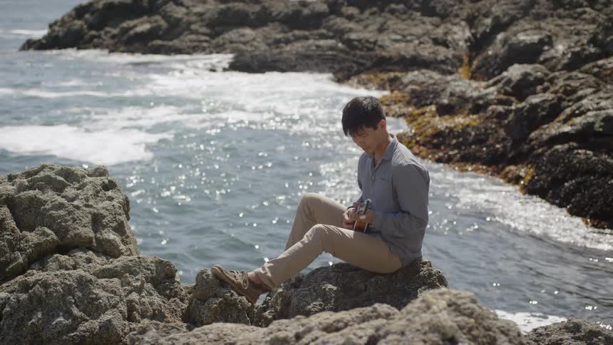A man sits on a rocky coast playing a small guitar