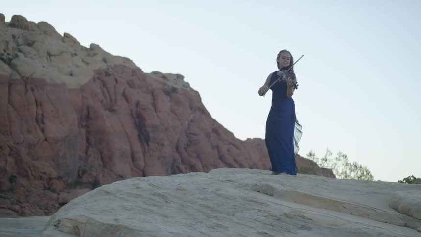 Blonde woman wearing a blue dress plays on the violin on the rock in the canyon
