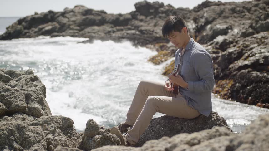 A man plays a ukulele on a rock near the crashing waves