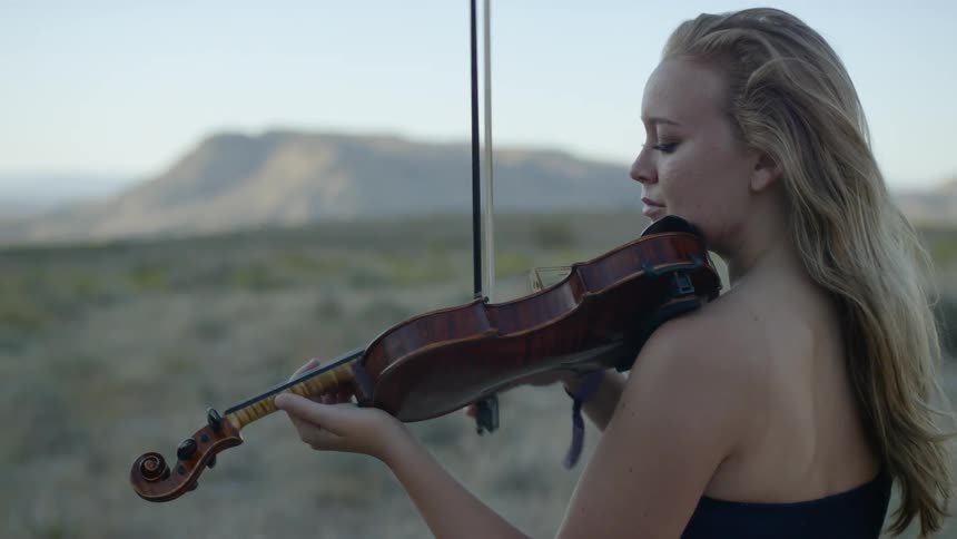 A woman standing outside playing the violin with the sky and mountains in the background