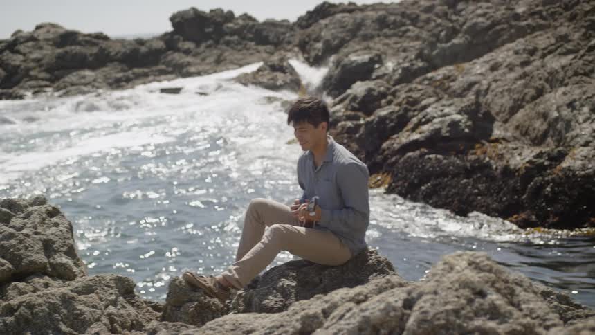 A man is sitting on a rock playing the ukulele as waves lap the shoreline nearby