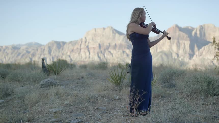 A white female violinist in a long blue dress uses a bow to play her instrument outside with mountains in the background