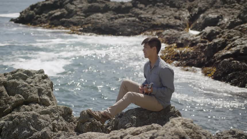 Man sits on rocks at shore strumming ukulele