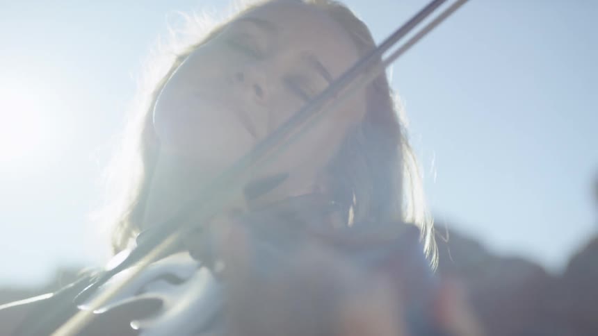 A blonde haired woman stands outside under the sunlight as she plays a violin