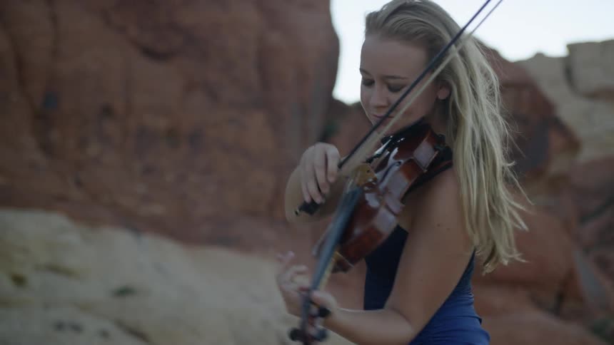 Blonde woman wearing a blue dress plays on the violin in the canyon