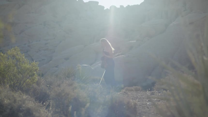 A woman walks through a grassy valley with a violin and bow