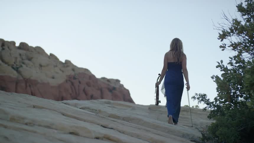 Woman in a formal dress walking barefoot carrying a violin in a canyon