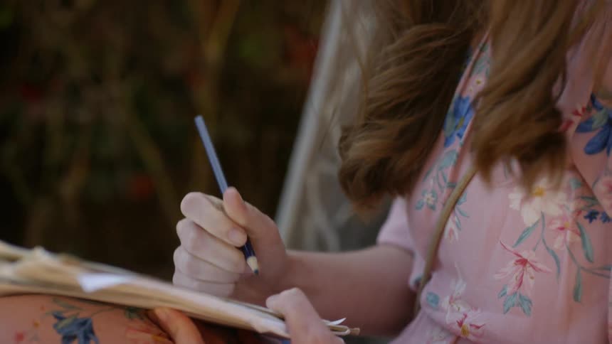 Woman in dress sits outside writing on notebook with pencil