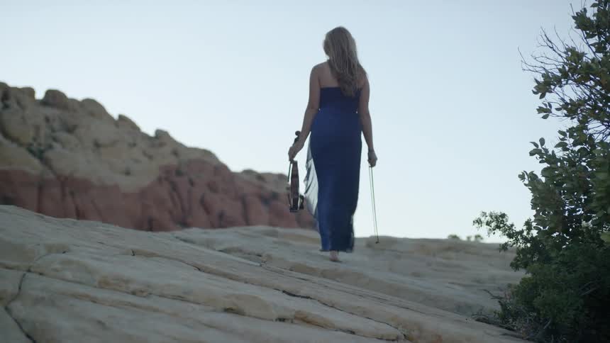 A woman walks an elevated rocky terrain carrying a violin and bow