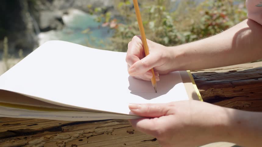 Woman leaning on railing near the ocean writes on notepad with pencil