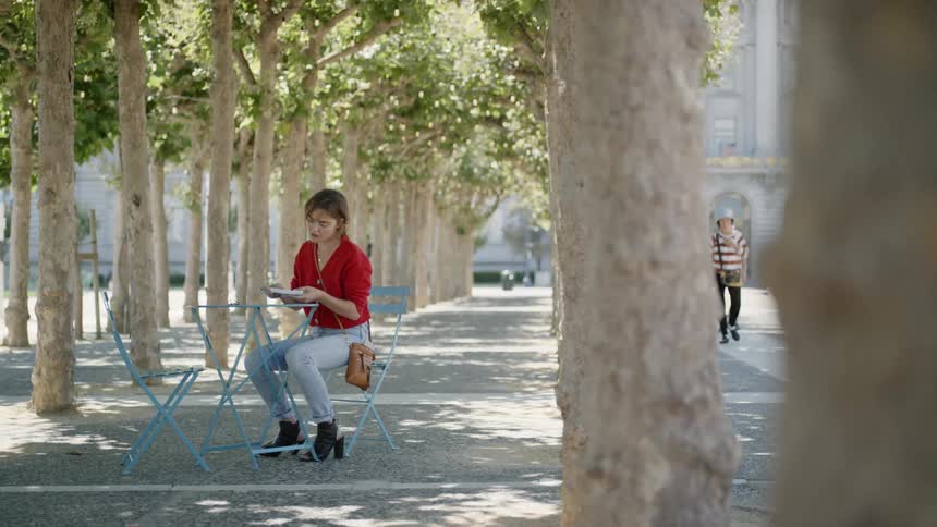 A young white woman sits at a small table outdoors and writes in a small notebook