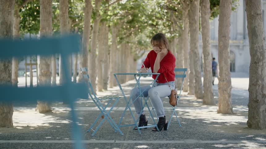 A young woman sits writing at a small table between two rows of trees in a citys public park