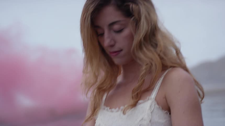 Pink smoke billows over ocean behind woman in sundress