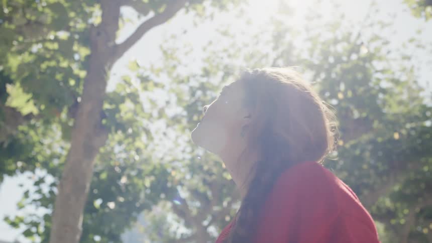 A woman is standing outdoors and looking up at the trees