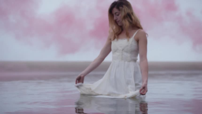 A woman in a white dress stands in a body of water and drags her skirt back and forth across the surface