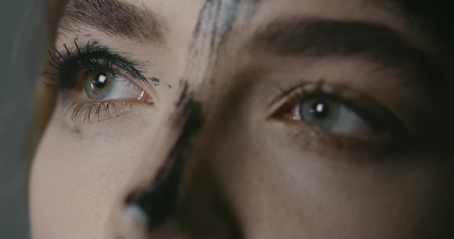 A woman with black paint across her nose and one eye slowly blinks