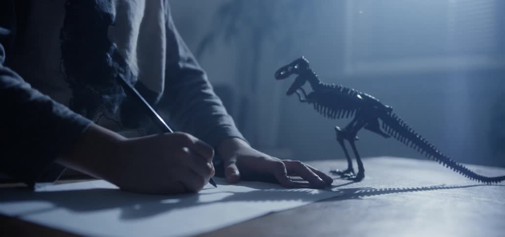 A child draws on a sheet of paper next to a dinosaur skeleton figurine