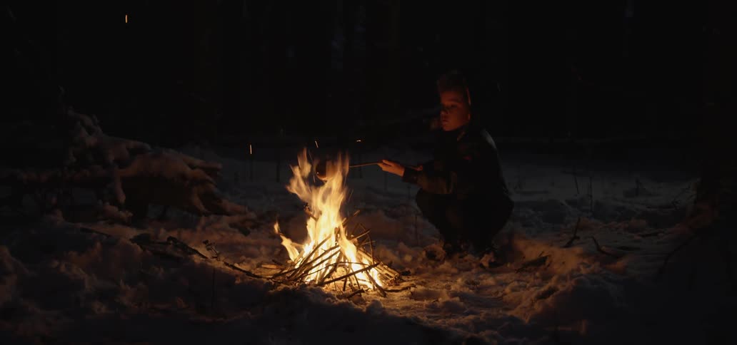 A child is cooking food over a fire in the snow at night