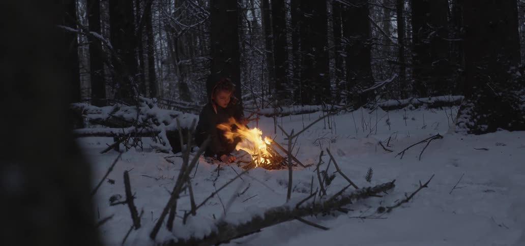 A boy adding twigs to a small campfire in the woods during the winter
