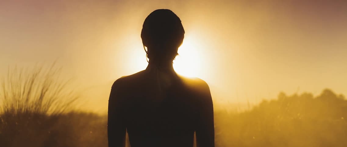 A naked woman rising into view as steam rolls around her and the sun sets in the distance