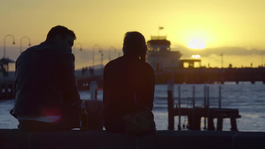 A man and woman sit and talk on a pier at sunset