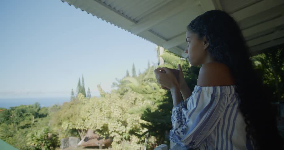 A young black woman with long curly hair sips her drink as she leans on the handrail of a balcony over a yard