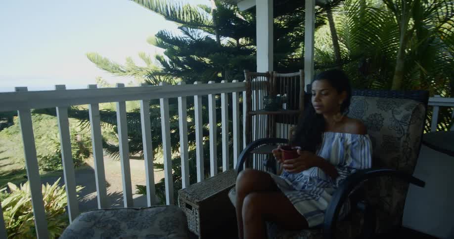 A woman walks onto a porch and sits down with a mug in hand
