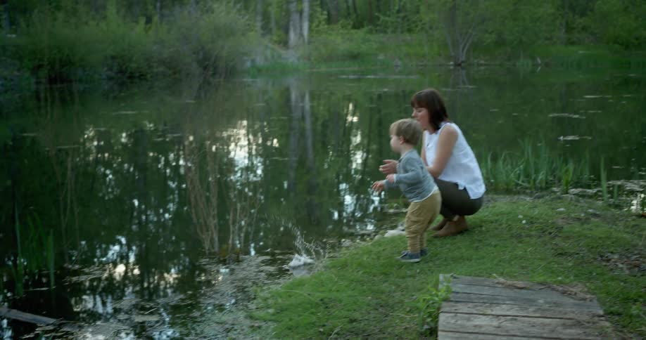 A toddler boy throws a rock into a lake while his mother crouches next to him