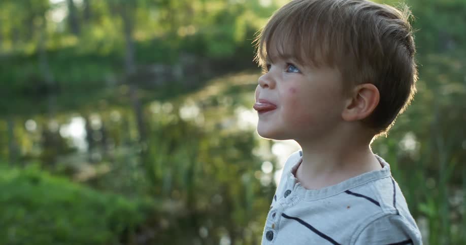 A young boy stands by a pond and sticks his tongue out