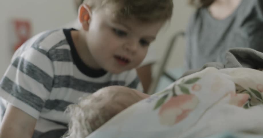 A toddler leans forward to look at a newborn baby in the hospital