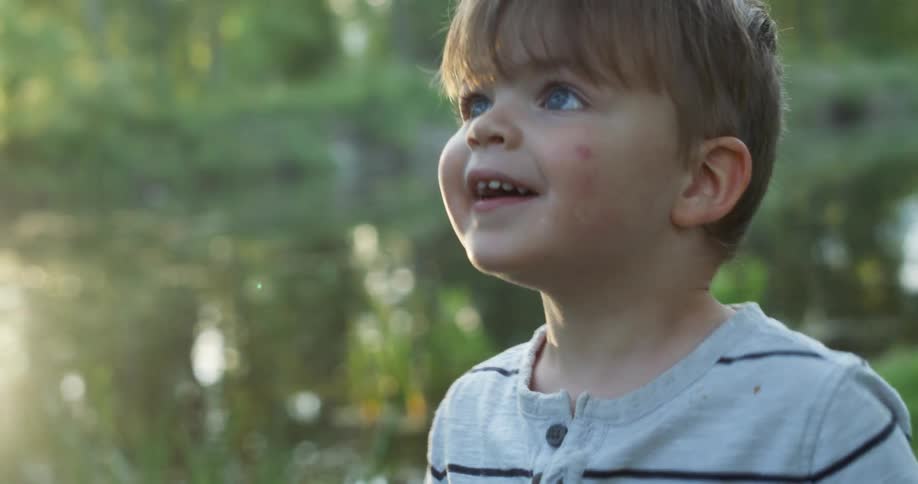 Blue eyed toddler boy looks up and smiles by the lake at sunset