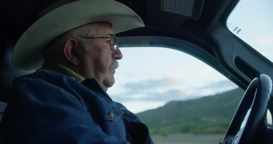 A white man wearing a cowboy hat and a denim jacket driving a vehicle