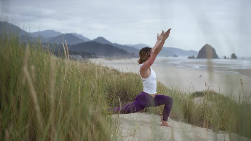 A woman stretches in a yoga pose in the sand next to a beach