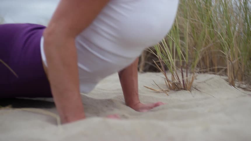 A woman in athletic clothes does yoga on a sandy beach with grass