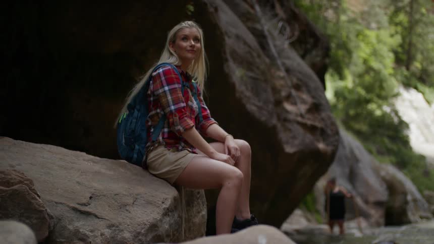 A teenager sits on a large rock near a stream