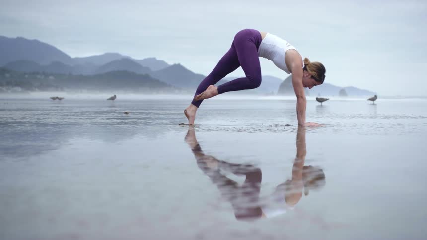 A woman stretches on the wet sand and lifts her leg up above her head in a yoga pose