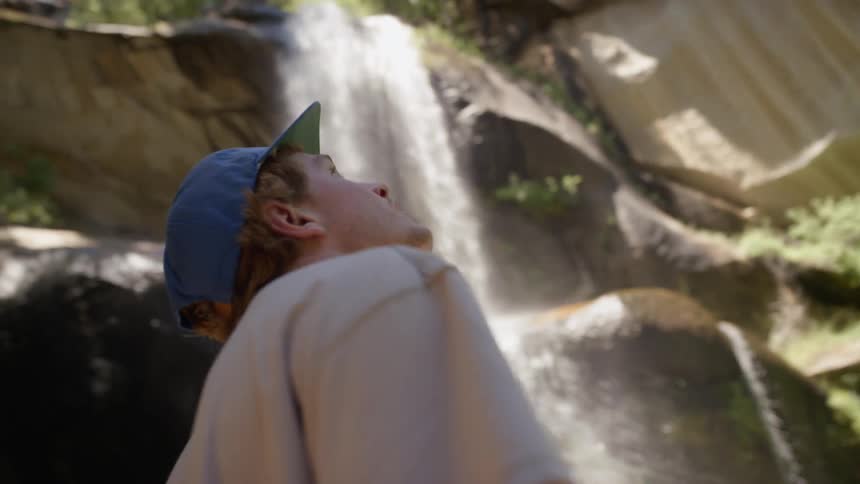 Man stands at foot of waterfall looking up toward sky