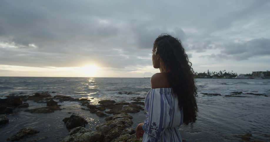 A woman with a flower in her hair stands on a beach at sunset