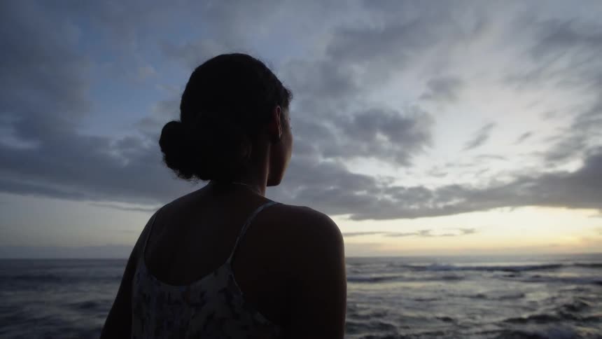 Brunette girl stands on the sand beach and looks at the sea at dusk