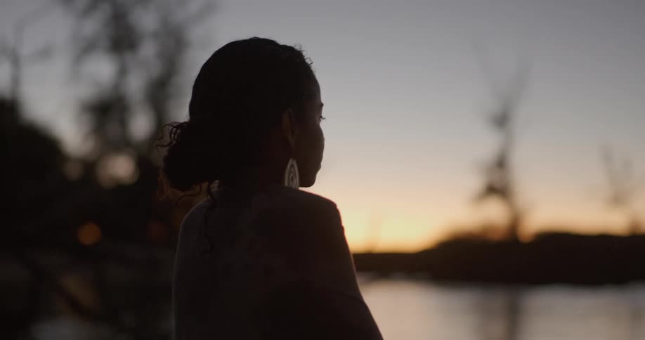 A woman stands near a lake at sunset