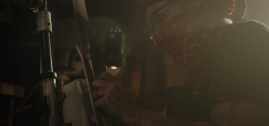 A person with a red bandana over their face sanding the handle of a knife with an electric sander