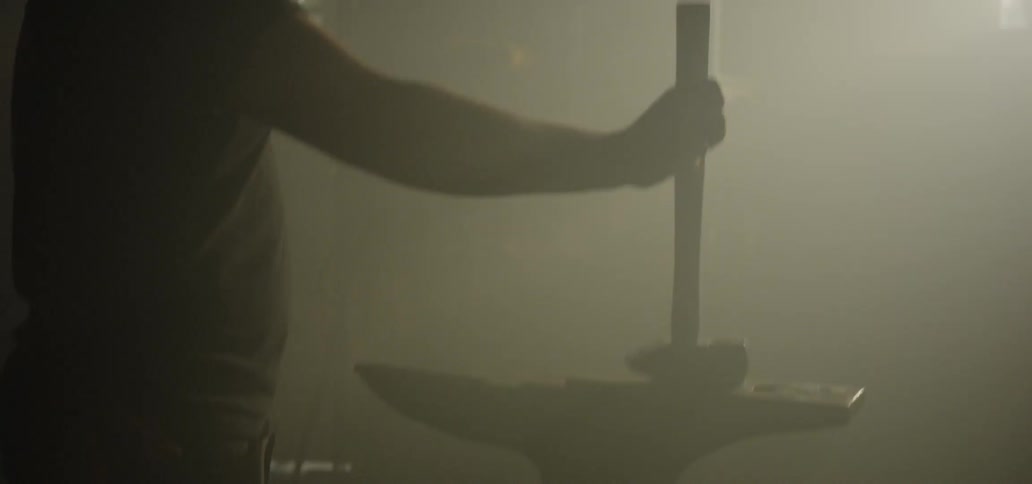 A man holding a blacksmith hammer rests it upon an anvil in a hazy room