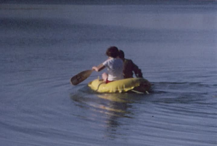 Two young boys leave the sandy shore of a lake paddling in a small rubber raft