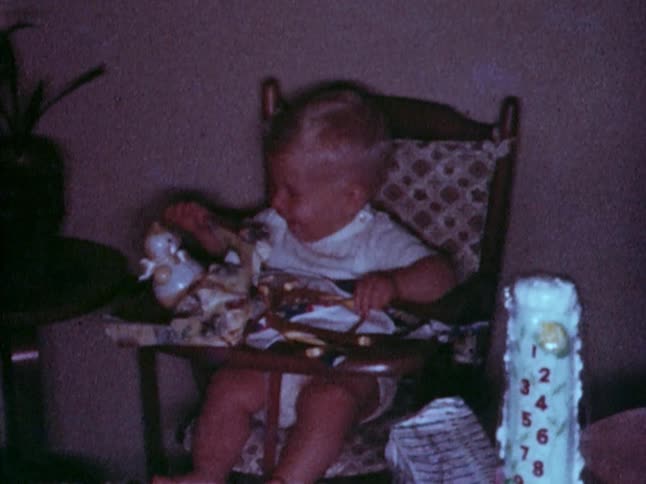 Old footage of a little boy sitting in a high chair smiling and touching a toy that is on top of wrapping paper on the chair's tray