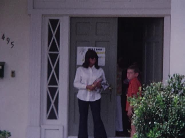 A blond boy runs across a yard as women come out of a house