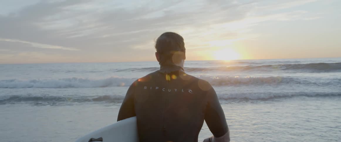 A man is carrying his surfboard toward the ocean as the sun rises